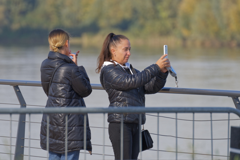 3 - Two individuals standing by a railing overlooking a body of water, with the person on the right taking a photograph with a mobile device